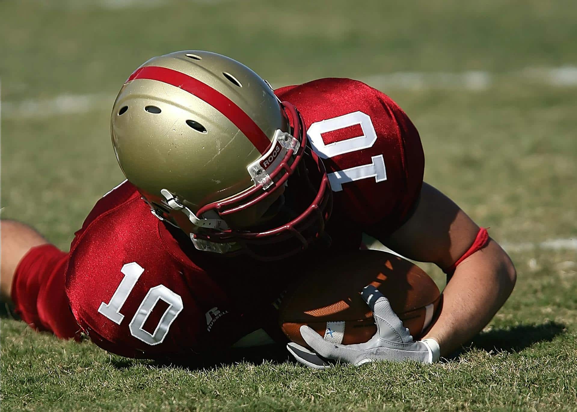 Man in red and gold football gear holds a football close while lying on the ground.