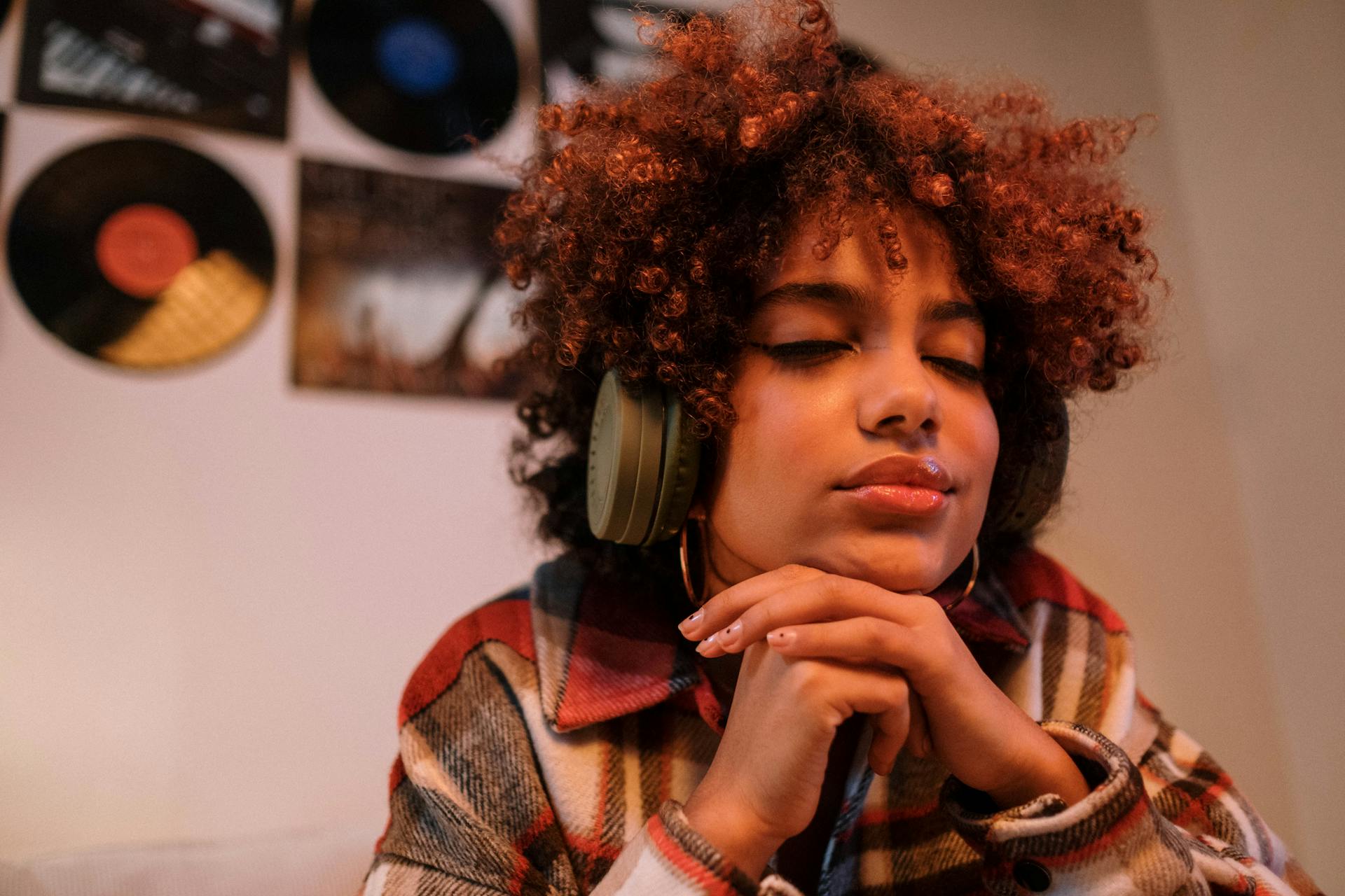Woman in chunky green headphones listening with her eyes closed with vinyl records hung on the wall in the background.